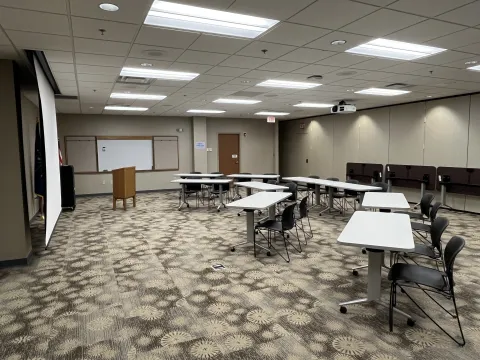 Jessie Thomas Meeting Room A with tables and chairs set up. The projector is also set up as well as a lectern. You can see the whiteboard on the back wall as well. 